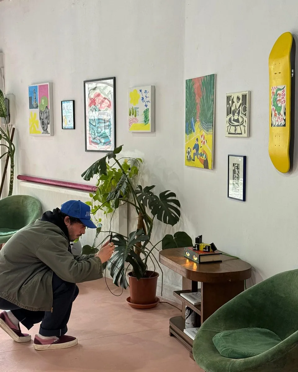 Expoziție în garaj: tablouri și o placă de skate pe pereți, cineva fotografiază lucrările